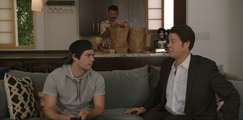Movie still from “Maximum Truth” (2023), directed by David Stassen – Two men sitting on a couch in front of a man with paper bags; Medium shot, Over the shoulder angle