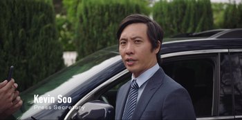 Movie still from “Maximum Truth” (2023), directed by David Stassen – A man in a suit and tie standing next to a car; Medium shot, Over the shoulder angle