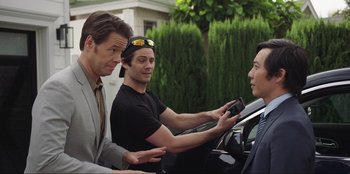 Movie still from “Maximum Truth” (2023), directed by David Stassen – Three men standing next to each other in front of a car; Medium shot, Over the shoulder angle