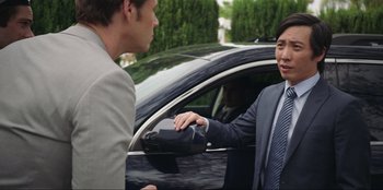 Movie still from “Maximum Truth” (2023), directed by David Stassen – A man in a business suit shaking hands with another man in front of a car; Medium shot, Over the shoulder angle