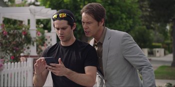 Movie still from “Maximum Truth” (2023), directed by David Stassen – Two men looking at a cell phone while standing next to each other; Medium shot, Over the shoulder angle
