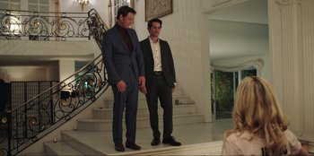 Movie still from “Maximum Truth” (2023), directed by David Stassen – A couple of men standing next to each other in front of a staircase; Wide shot, Over the shoulder angle