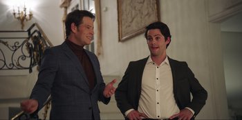 Movie still from “Maximum Truth” (2023), directed by David Stassen – Two men in suits talking to each other in front of a wall; Medium shot, Over the shoulder angle