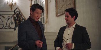 Movie still from “Maximum Truth” (2023), directed by David Stassen – Two men in suits talking to each other in front of a staircase; Medium shot, Over the shoulder angle