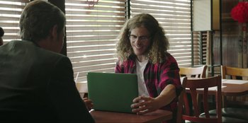 Movie still from “Maximum Truth” (2023), directed by David Stassen – A man sitting in front of a laptop computer at a table; Medium shot, Over the shoulder angle