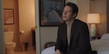 Movie still from “Maximum Truth” (2023), directed by David Stassen – A man in a suit sitting in a chair in front of a painting; Medium shot, Over the shoulder angle