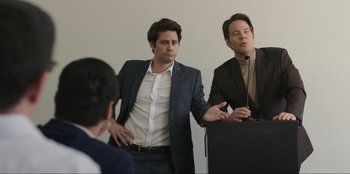 Movie still from “Maximum Truth” (2023), directed by David Stassen – Two men in business suits are standing in front of a podium; Medium shot, Over the shoulder angle