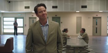 Movie still from “Maximum Truth” (2023), directed by David Stassen – A man in a suit standing next to a table; Medium shot, Over the shoulder angle