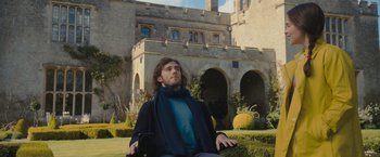 Movie still from “Me Before You” (2016), directed by Thea Sharrock – A man sitting in a chair in front of a building; Medium shot, Low angle