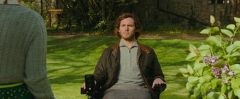 Movie still from “Me Before You” (2016), directed by Thea Sharrock – A man sitting in a chair in the grass; Medium shot, Over the shoulder angle