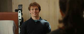 Movie still from “Me Before You” (2016), directed by Thea Sharrock – A young man smiling while standing in front of a wooden wall; Close Up shot, Over the shoulder angle