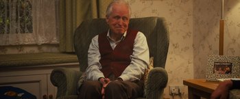 Movie still from “Me Before You” (2016), directed by Thea Sharrock – An older man sitting in a chair with his hands crossed; Medium shot, Over the shoulder angle