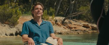 Movie still from “Me Before You” (2016), directed by Thea Sharrock – A man sitting on the ground near a body of water; Medium shot, Over the shoulder angle