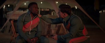 Movie still from “Me Time” (2022), directed by John Hamburg – Two men sitting on the ground one of which is holding a cell phone; Medium shot, Over the shoulder angle