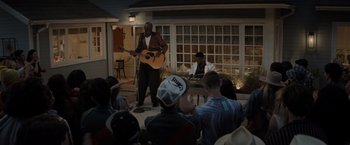 Movie still from “Me Time” (2022), directed by John Hamburg – A man playing a guitar in front of a group of people; Wide shot, Over the shoulder angle