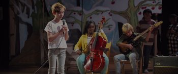 Movie still from “Me Time” (2022), directed by John Hamburg – A group of people sitting on a stage playing instruments; Medium shot, Over the shoulder angle