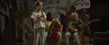 Movie still from “Me Time” (2022), directed by John Hamburg – A young girl holding a microphone while standing in front of a group of young people; Medium shot, Over the shoulder angle