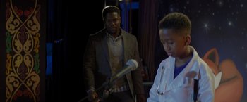 Movie still from “Me Time” (2022), directed by John Hamburg – A man holding a microphone in front of another man in a white lab coat; Medium shot, Over the shoulder angle