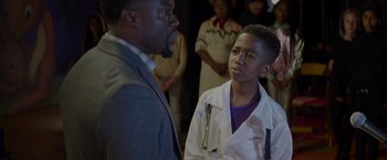 Movie still from “Me Time” (2022), directed by John Hamburg – A man and a young boy in a white lab coat talking to each other in front of a crowd; Medium shot, Over the shoulder angle