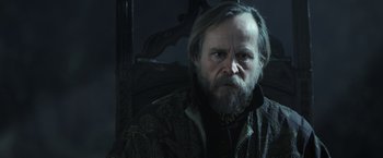 Movie still from “Medieval” (2022), directed by Petr Jákl – A man with long hair and a beard sitting in a chair; Close Up shot, Over the shoulder angle