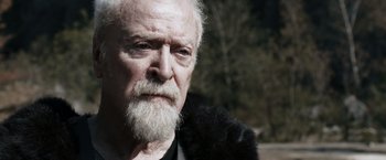Movie still from “Medieval” (2022), directed by Petr Jákl – An old man with a beard and a black jacket; Close Up shot, Low angle