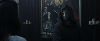 Movie still from “Medieval” (2022), directed by Petr Jákl – A man wearing a black hooded sweatshirt sitting in front of a painting; Medium shot, Low angle