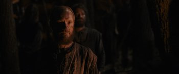 Movie still from “Medieval” (2022), directed by Petr Jákl – A man with a long beard and a red beard is standing in a dark room; Medium shot, Over the shoulder angle