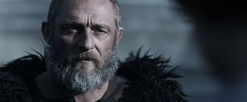 Movie still from “Medieval” (2022), directed by Petr Jákl – An old man with a beard and a black jacket; Close Up shot, Low angle