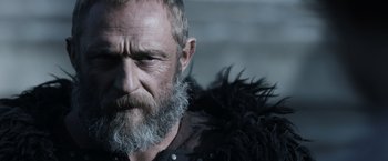 Movie still from “Medieval” (2022), directed by Petr Jákl – An older man with a beard and a black jacket; Close Up shot, Low angle