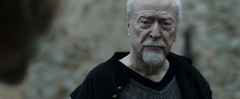 Movie still from “Medieval” (2022), directed by Petr Jákl – An older man with a beard and mustache wearing a black shirt; Close Up shot, Over the shoulder angle