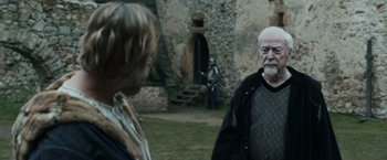 Movie still from “Medieval” (2022), directed by Petr Jákl – A man and a woman standing in front of a stone building; Medium shot, Over the shoulder angle