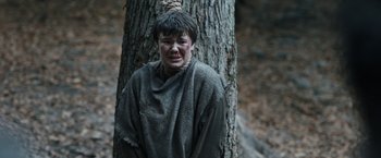 Movie still from “Medieval” (2022), directed by Petr Jákl – A young boy in a hooded blanket hanging from a tree; Medium shot, Low angle