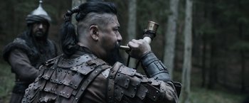 Movie still from “Medieval” (2022), directed by Petr Jákl – A man with a long beard holding an object in his mouth; Close Up shot, Over the shoulder angle