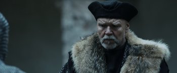Movie still from “Medieval” (2022), directed by Petr Jákl – An old man with a beard wearing a fur coat and a hat; Close Up shot, Over the shoulder angle