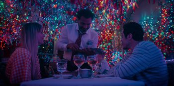 Movie still from “Meet Cute” (2022), directed by Alex Lehmann – A man in a tie is standing at a table with three glasses of wine; Medium shot, Over the shoulder angle