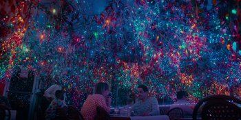 Movie still from “Meet Cute” (2022), directed by Alex Lehmann – Two people are sitting at a table in front of a bunch of christmas lights; Extreme Wide shot, Over the shoulder angle