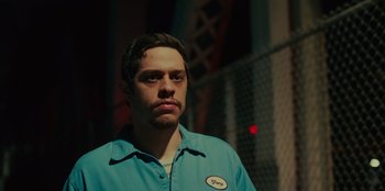 Movie still from “Meet Cute” (2022), directed by Alex Lehmann – A man in a blue shirt standing in front of a chain link fence; Close Up shot, Low angle