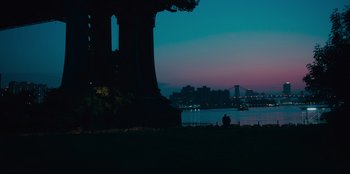 Movie still from “Meet Cute” (2022), directed by Alex Lehmann – Two people sitting on a bench near a body of water; Extreme Wide shot, Low angle