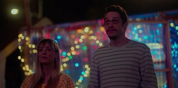 Movie still from “Meet Cute” (2022), directed by Alex Lehmann – A man and a woman standing next to each other in front of lights; Medium shot, Over the shoulder angle