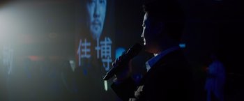 Movie still from “Meg 2: The Trench” (2023), directed by Ben Wheatley – A man holding a microphone in front of a picture of a man; Close Up shot, Over the shoulder angle