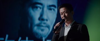 Movie still from “Meg 2: The Trench” (2023), directed by Ben Wheatley – A man holding a microphone while standing in front of a picture of a man; Close Up shot, Over the shoulder angle