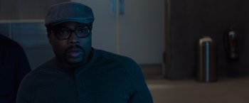 Movie still from “Meg 2: The Trench” (2023), directed by Ben Wheatley – A man wearing glasses and a hat sitting in a room; Close Up shot, Over the shoulder angle
