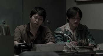 Movie still from “Memories of Murder” (2003), directed by Bong Joon Ho – Two asian men sitting at a table writing on a piece of paper; Medium shot, High angle