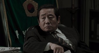 Movie still from “Memories of Murder” (2003), directed by Bong Joon Ho – An older man in a suit and tie sitting in an office chair; Close Up shot, Low angle