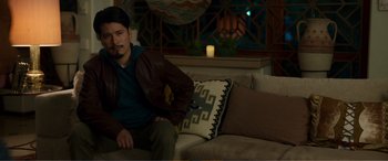 Movie still from “Memory” (2022), directed by Martin Campbell – A man sitting on top of a couch in a living room; Medium shot, Over the shoulder angle