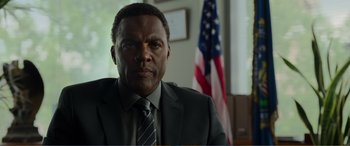 Movie still from “Memory” (2022), directed by Martin Campbell – A man wearing a suit and tie in front of a flag; Close Up shot, Low angle