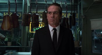 Movie still from “Men in Black” (1997), directed by Barry Sonnenfeld – A man in a suit and tie standing in front of guitars; Medium shot, Low angle