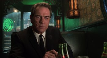 Movie still from “Men in Black” (1997), directed by Barry Sonnenfeld – A man in a suit and tie sitting at a table with a bottle; Close Up shot, Over the shoulder angle