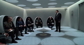 Movie still from “Men in Black” (1997), directed by Barry Sonnenfeld – A group of people sitting in a room with a man standing in front of them; Wide shot, High angle