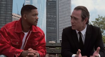 Movie still from “Men in Black” (1997), directed by Barry Sonnenfeld – A man in a red jacket and a man in a black suit talking; Medium shot, Over the shoulder angle